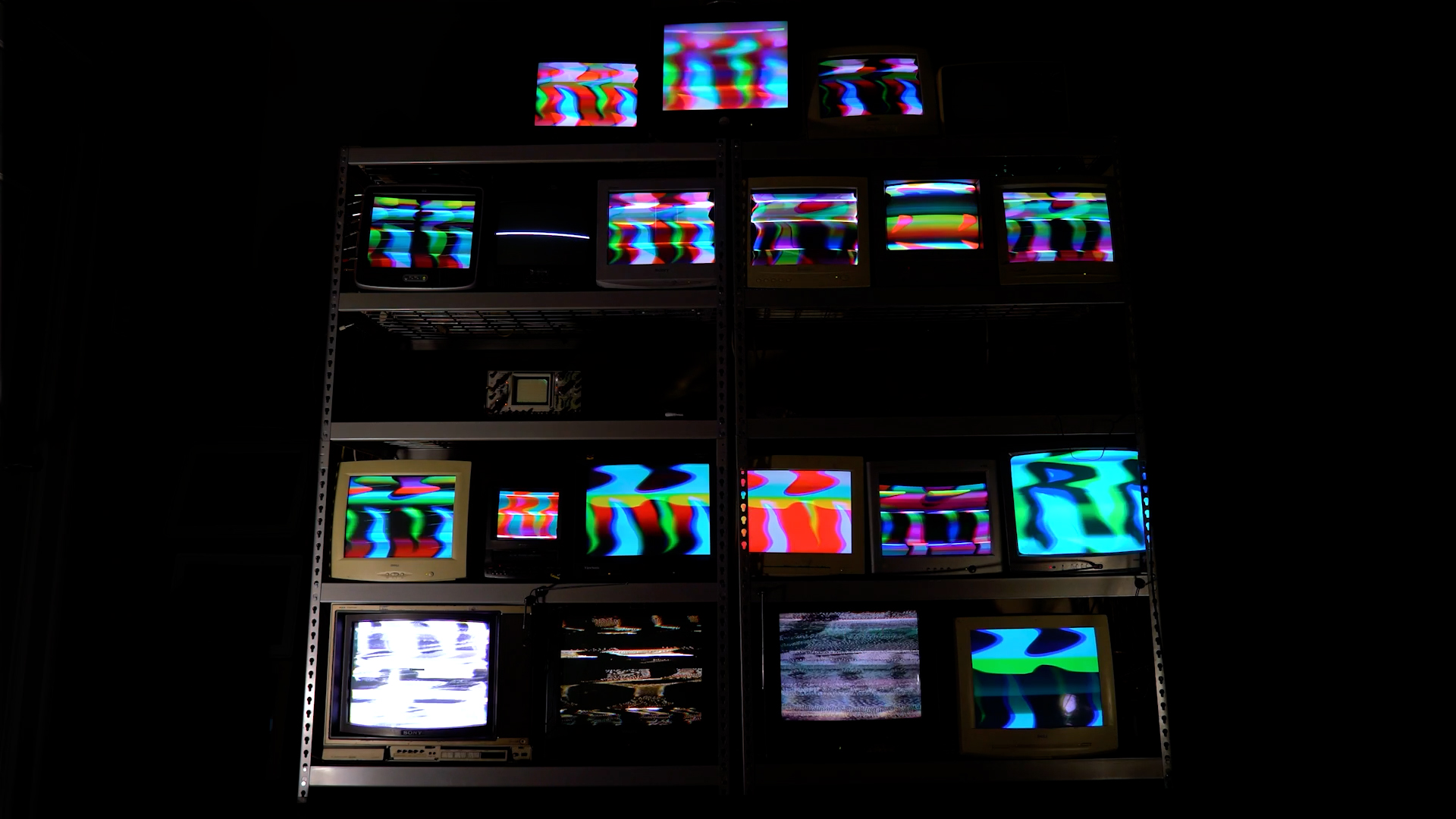 Still from Cathode Ray Algorithms showing a stack of 19 TV and VGA CRT monitors and an oscilloscope, all showing bright colors with electromagnetically manipulated shapes