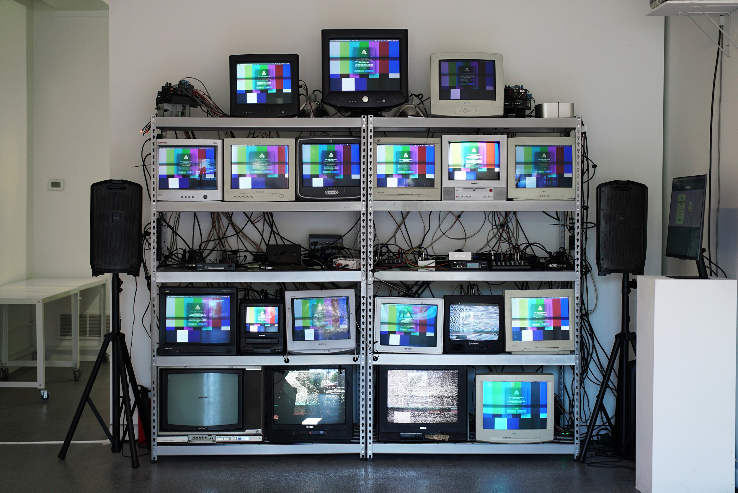19 CRT television and computer monitors on a metal shelf with color bars and an error promp. Speakers on stands are on either side of the shelf, and on the right, a flatscreen monitor is on a pedastal.