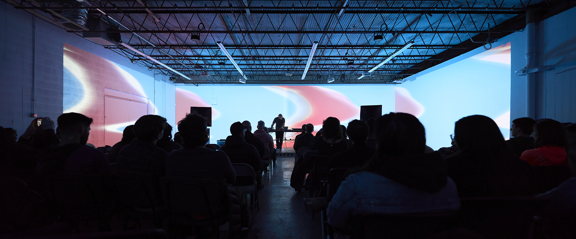 Looking through a crowd toward a stage with James Connolly performing with three walls of projected video synthesis visuals visuals around him.