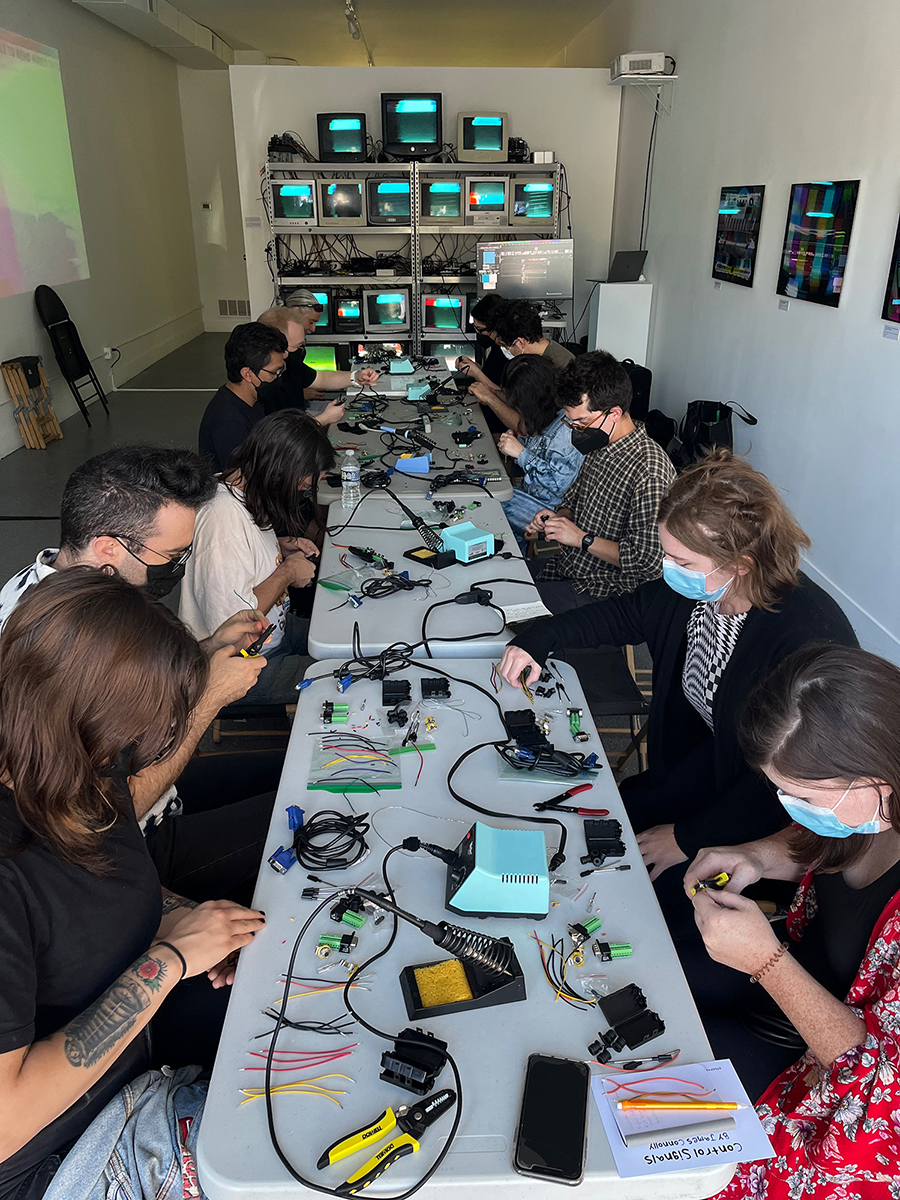 12 individuals participating in a DIY video synthesis workshop. Soldring irons and electronic components are on the tables, and a stack of monitors is seen in the background from James Connolly's Control Signals exhibition.