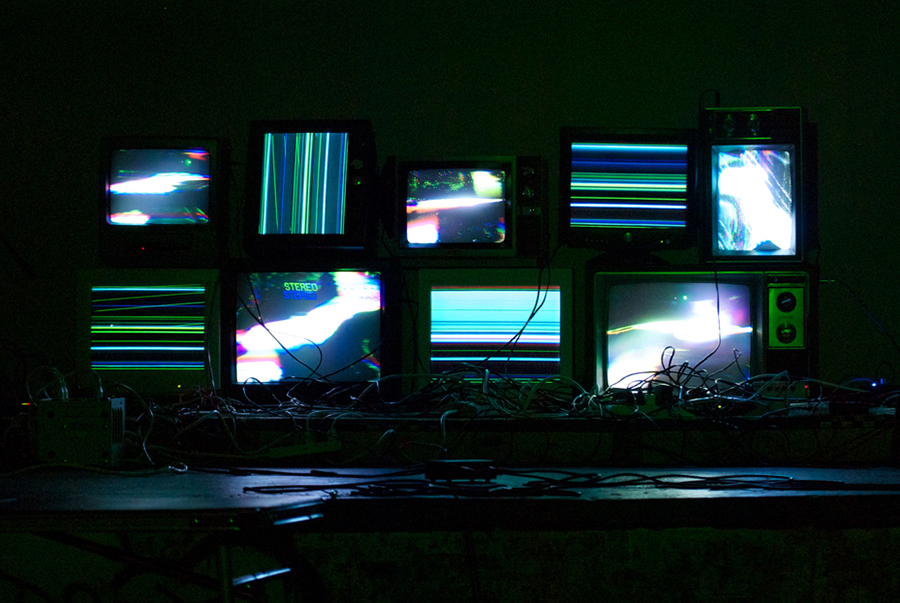 9 CRT television and computer monitos showing glitchy video with a large amount of cables surrounding
